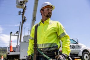 Lineworker wearing high-visibility flame-resistant (FR) shirt and PPE at an active utility job site with service trucks and power lines.