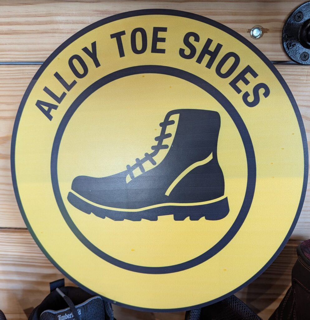 Circular “Alloy Toe Shoes” sign with a boot icon displayed on a wooden wall.