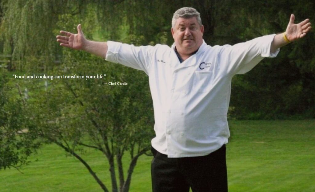 Chef Cardie Mortimer stands outdoors with open arms, wearing a white coat embroidered chef coat from Work Hard Dress Right, embodying his message that food and cooking can transform lives.