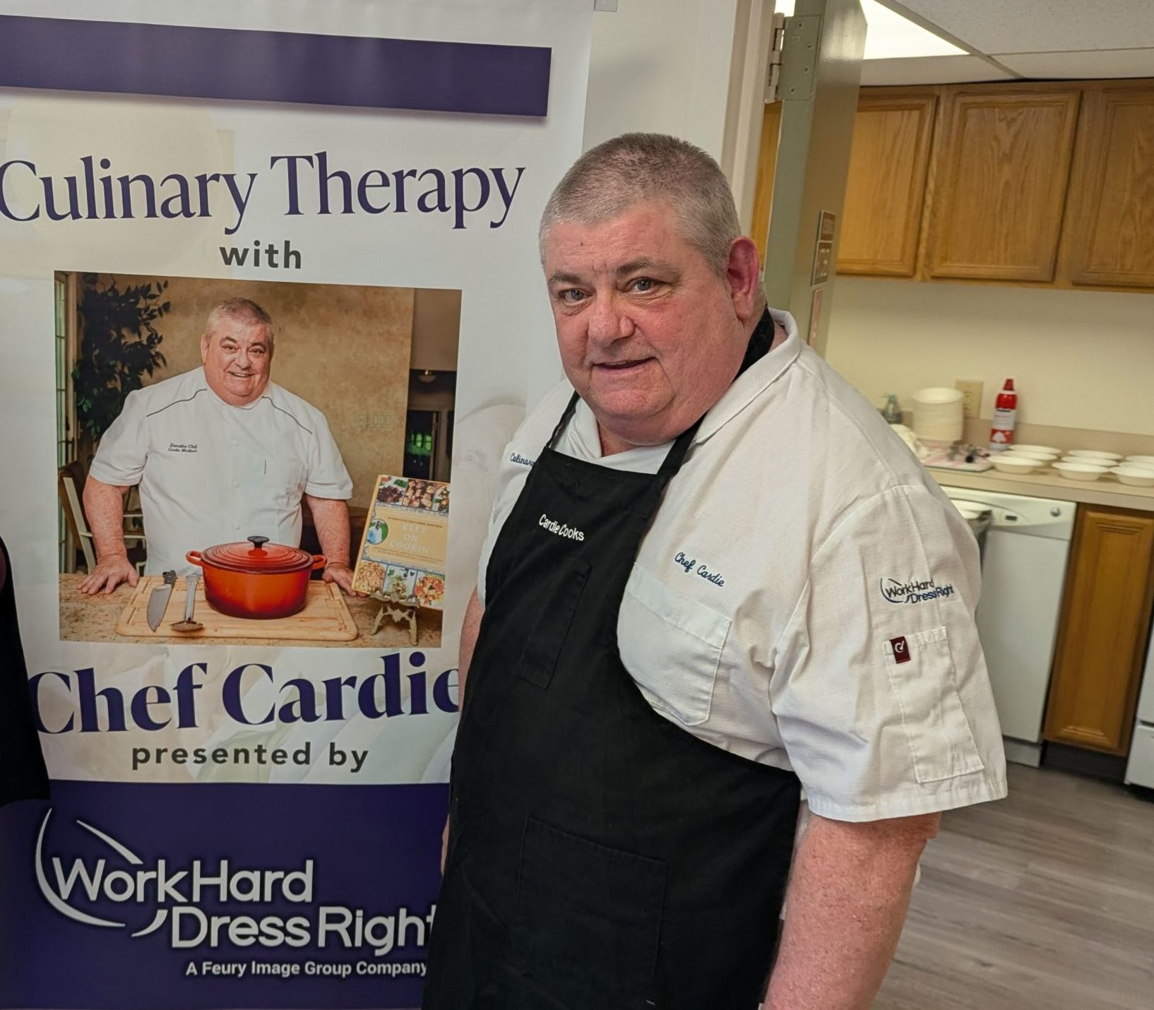 PXL_20251105_174600524 Chef Cardie Mortimer stands beside a Culinary Therapy banner presented by Work Hard Dress Right, wearing his custom embroidered chef coat and apron.
