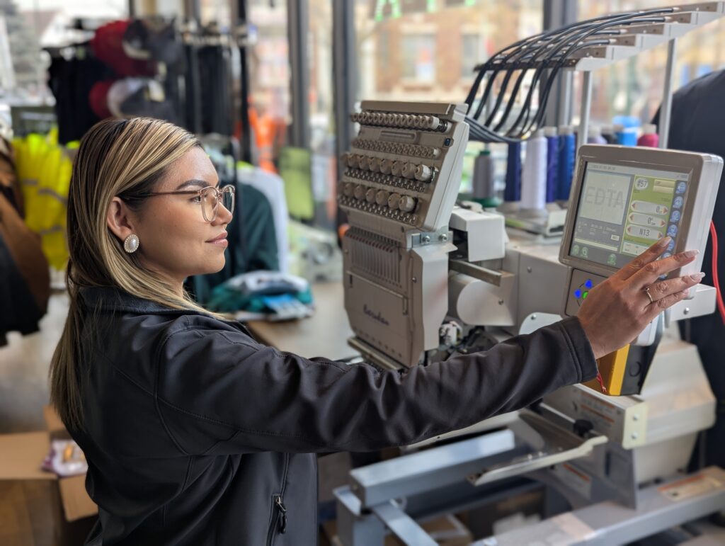 Erica operating an embroidery machine inside the Work Hard Dress Right store in Newark.